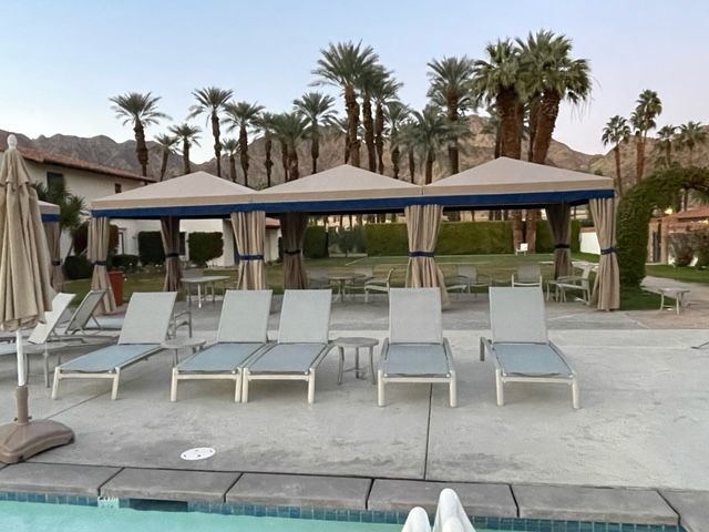 Lounge chairs by a pool under cabanas, palm trees, and mountain backdrop.