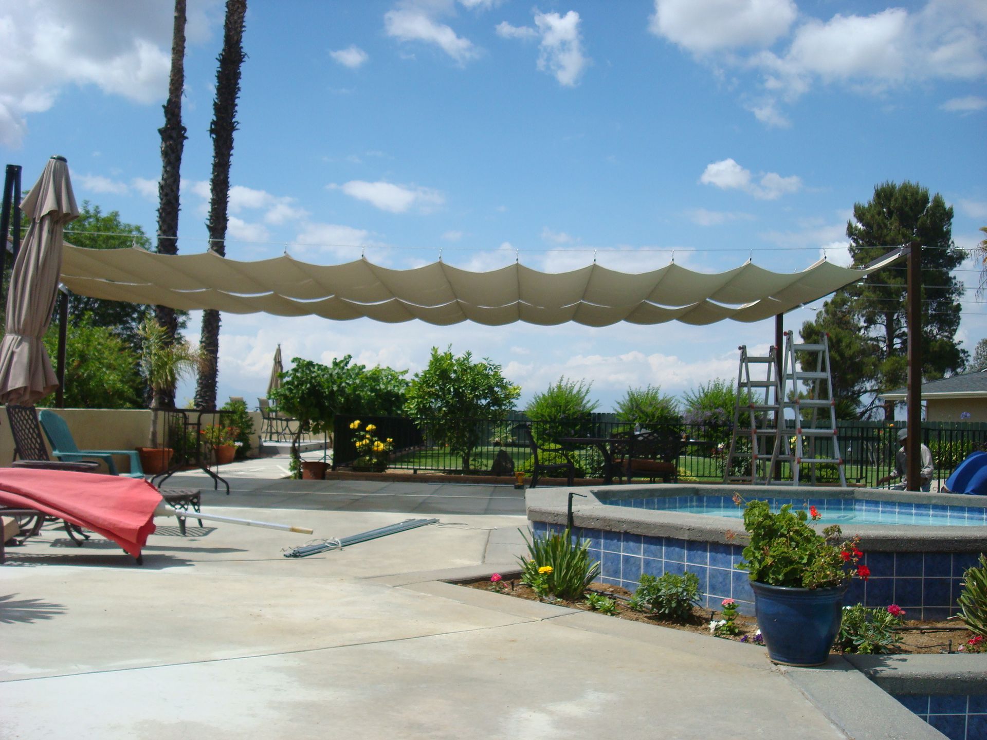 Shade structure over a pool and patio, sunny day. Brown fabric canopy, blue pool water, green plants.