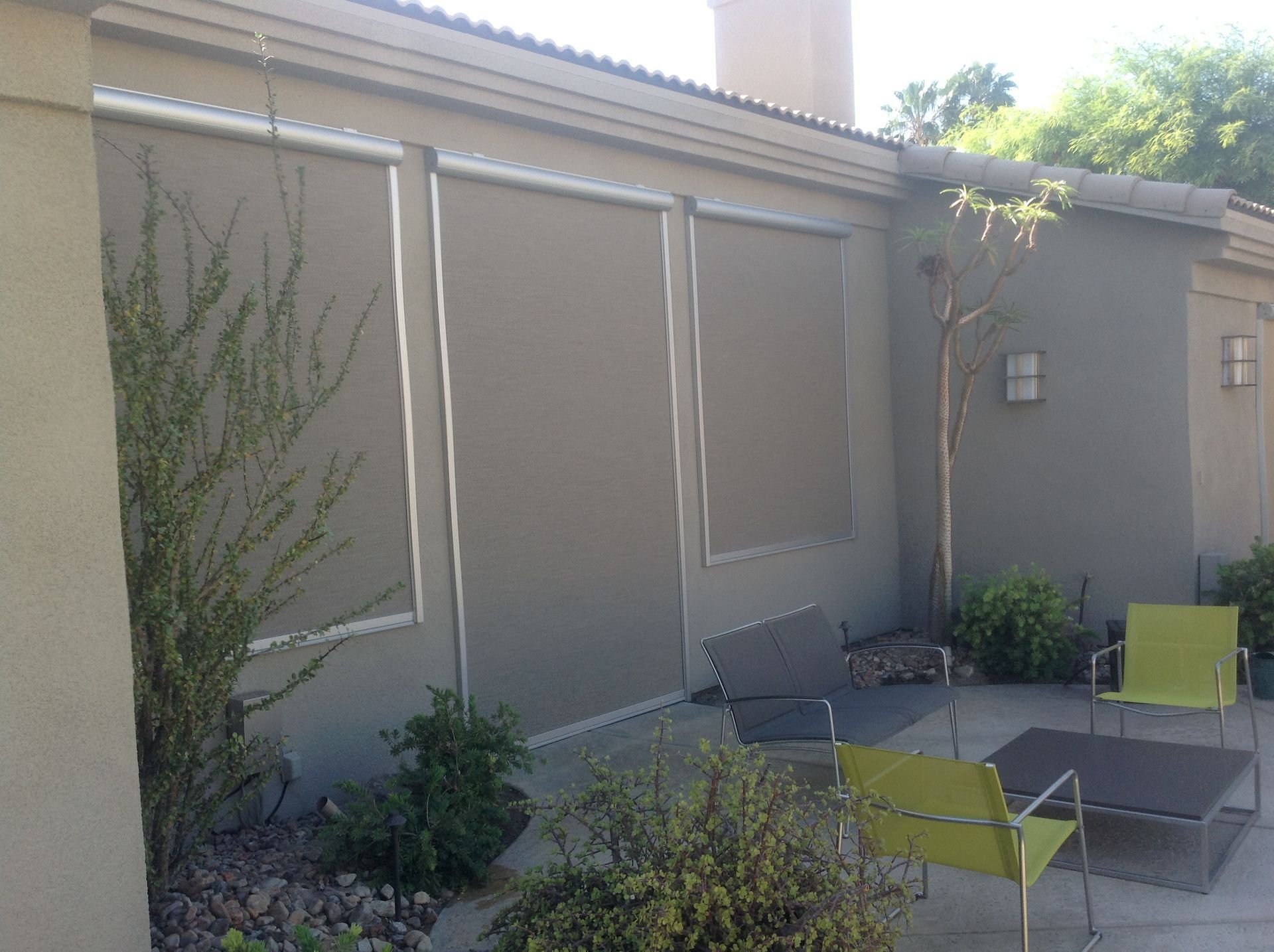 Outdoor patio with tan sun shades, seating, and greenery against a stucco wall.