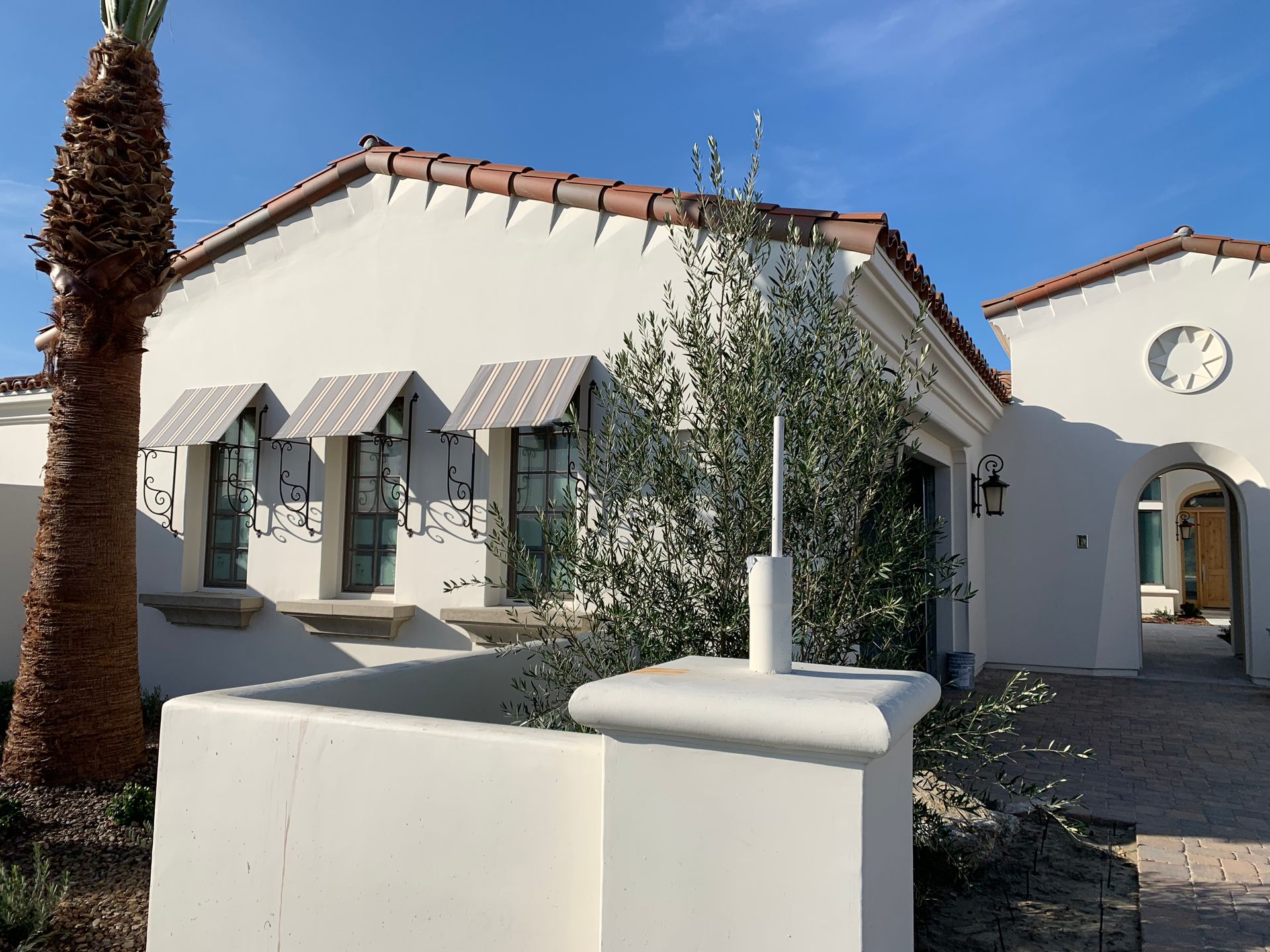 White stucco home with awnings, red tile roof, palm tree, and olive tree against a blue sky.