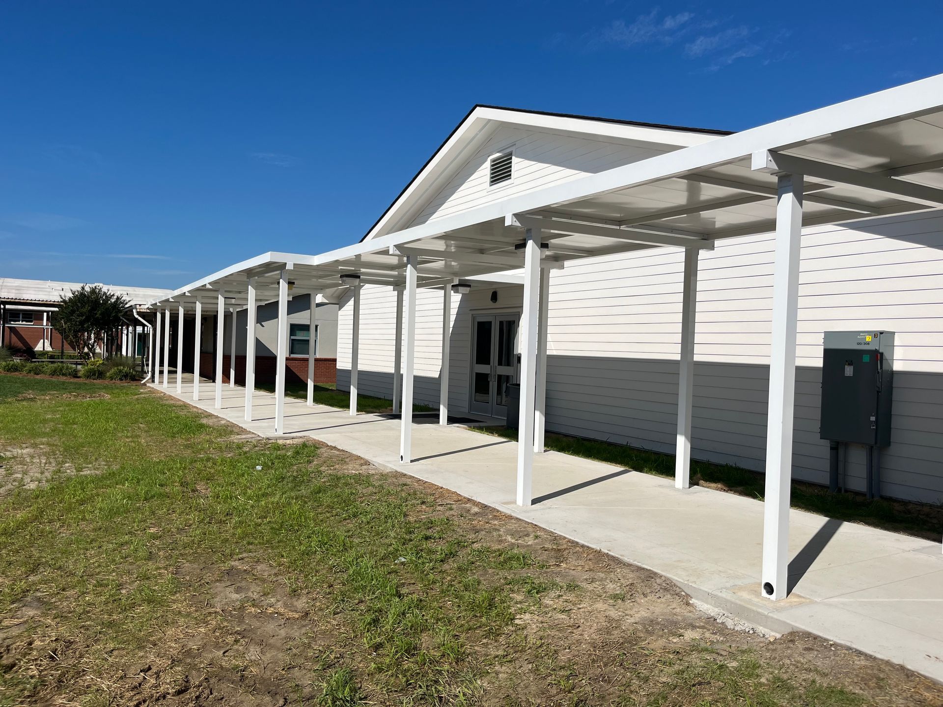 White canopy over a sidewalk beside a building with a door. Green grass and blue sky.