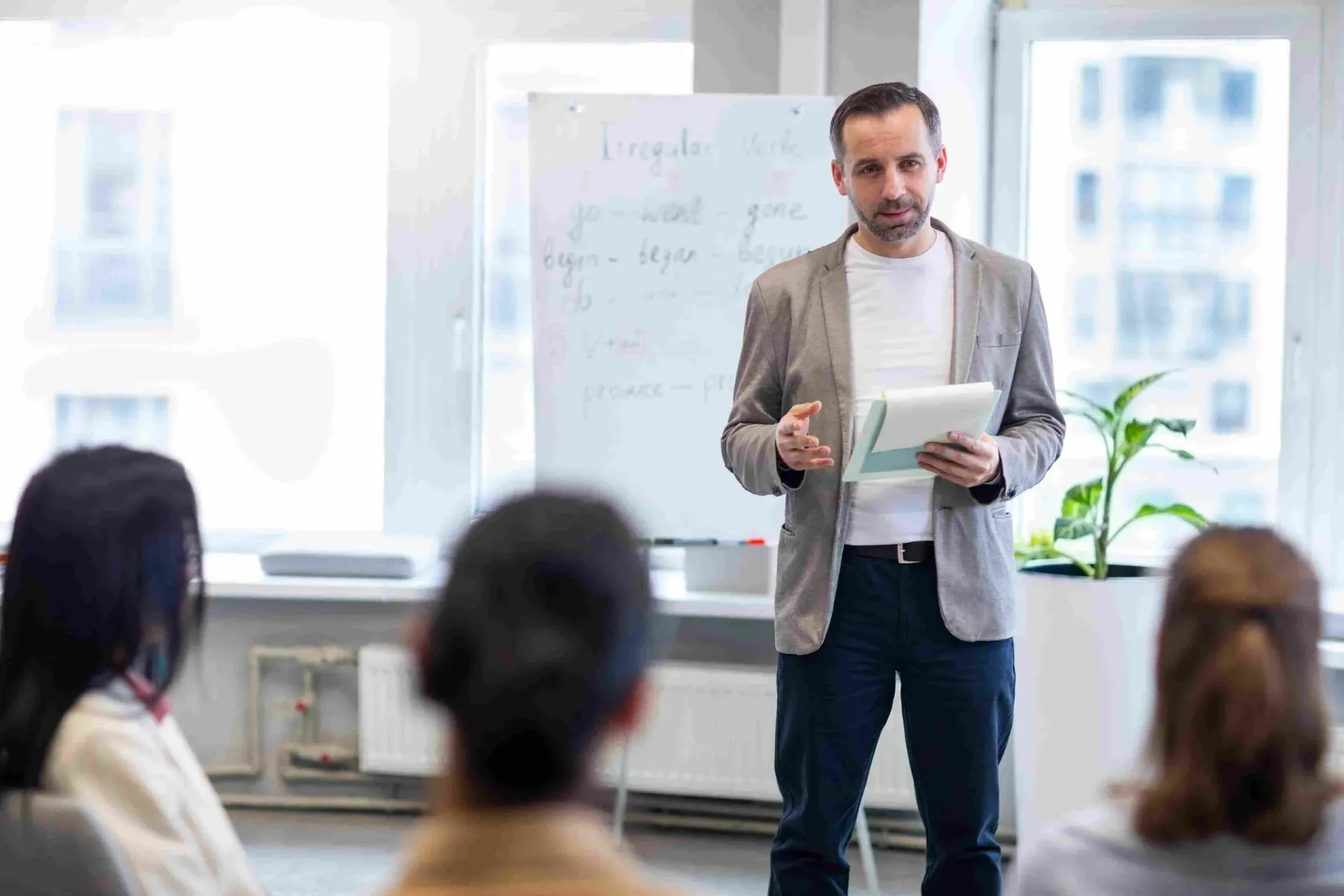 Man presenting in a bright office to seated audience, holding notes beside a whiteboard and plant.