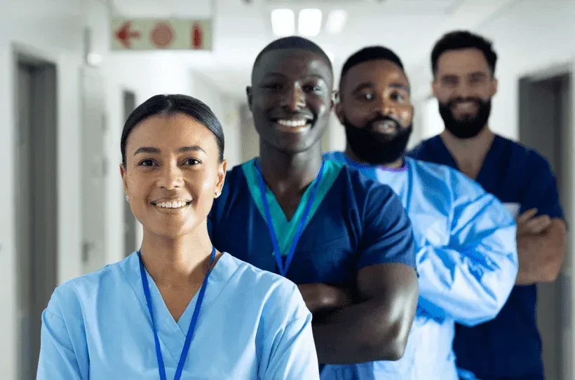 Four healthcare workers in blue scrubs smiling in a hospital hallway