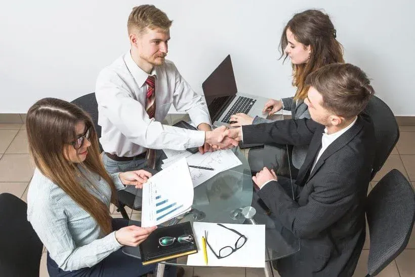 Business team in a meeting, shaking hands around a desk with a laptop and documents