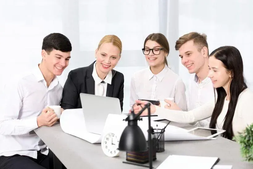 Five coworkers smiling around a laptop in a bright office meeting room