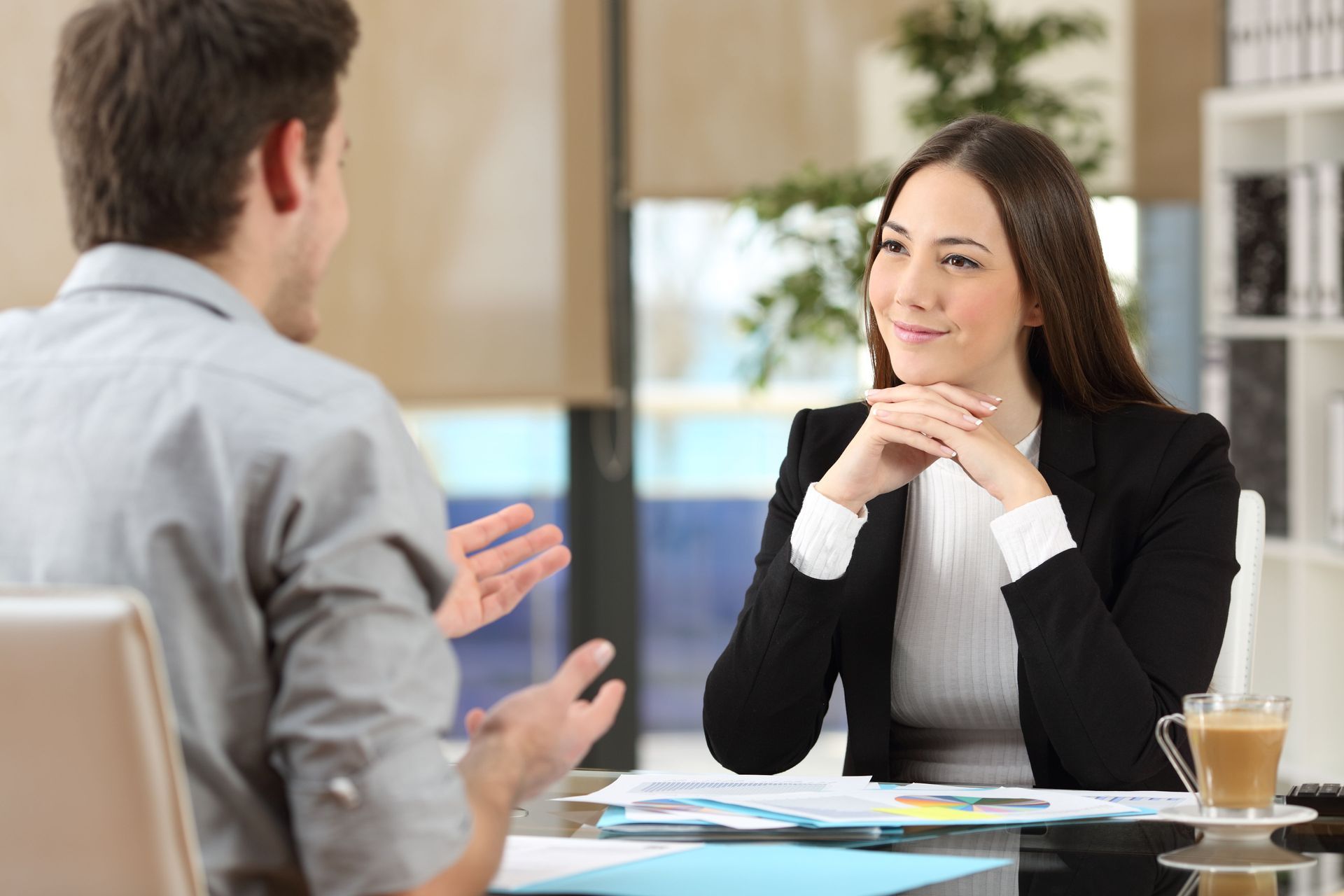 Two people talking across a desk in a bright office, with papers and a coffee cup nearby.