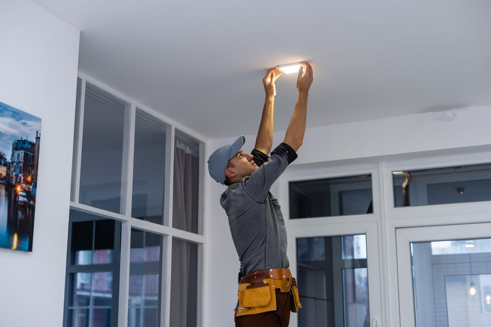Worker replacing a ceiling light in a bright room, standing on a ladder with tools on his belt