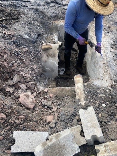 Worker in blue shirt and hat digging in a rocky excavation site with broken concrete blocks.