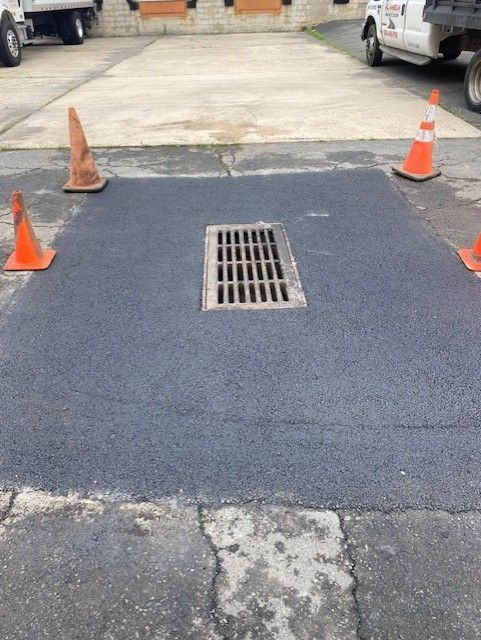 Newly paved asphalt around a storm drain, marked by orange traffic cones in a parking lot