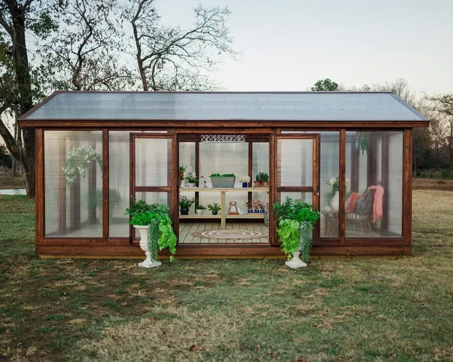 Wooden greenhouse with open doors, potted plants, and interior shelving on a grassy lawn.