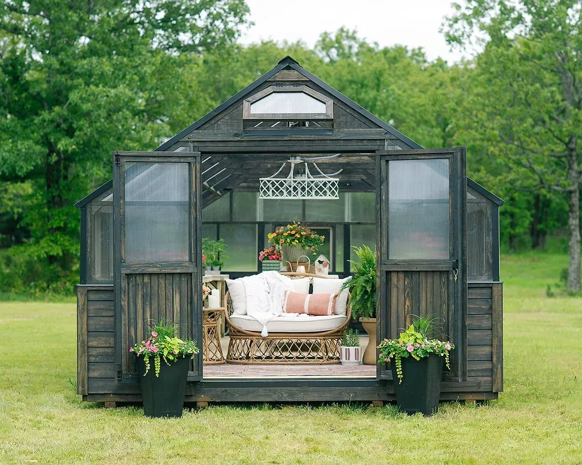 Black greenhouse with open doors, furnished with a sofa, plants, and a chandelier, set in a green field.