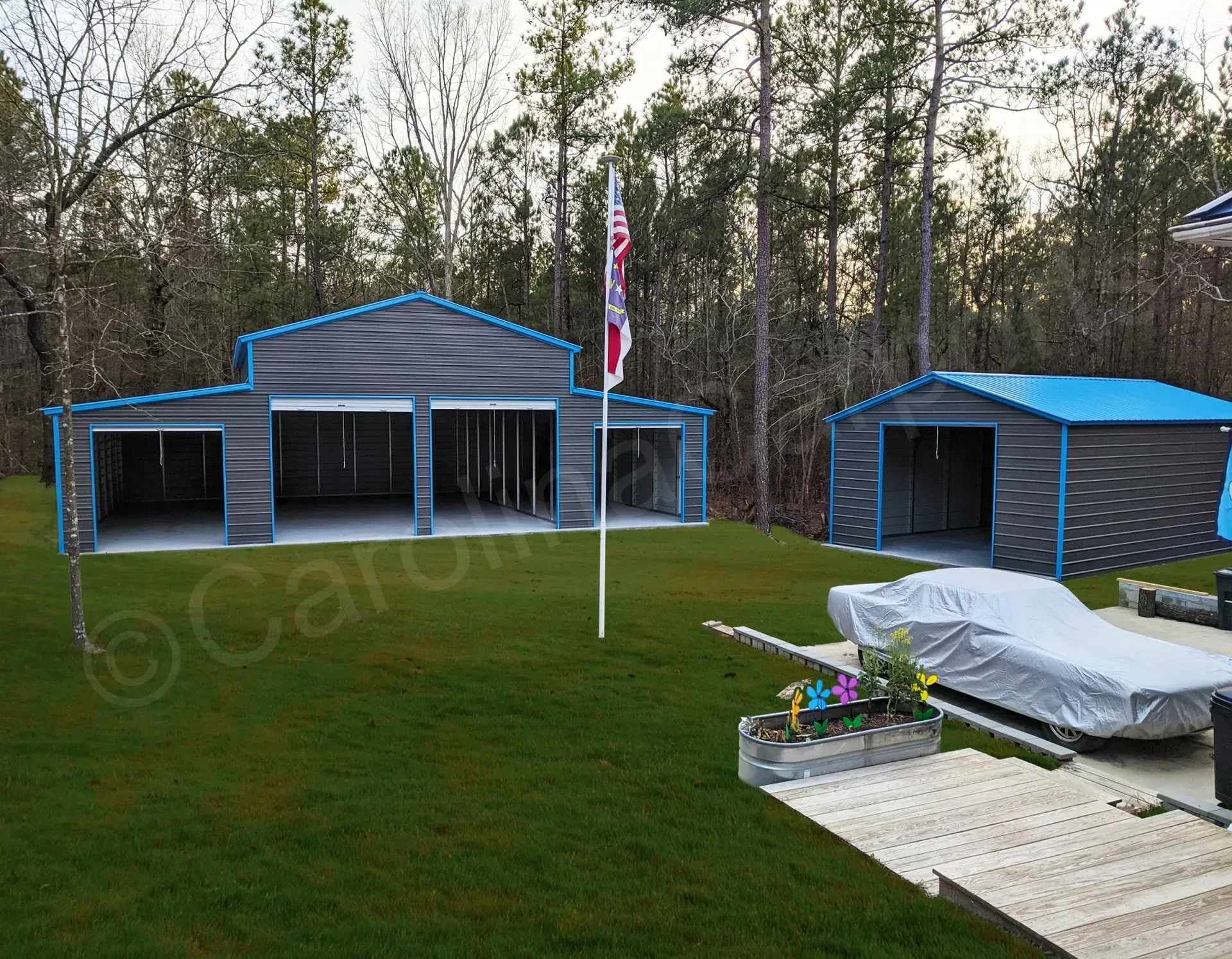 A gray metal garage and shed with blue trim on a green lawn with a covered car, flag pole, and trees.