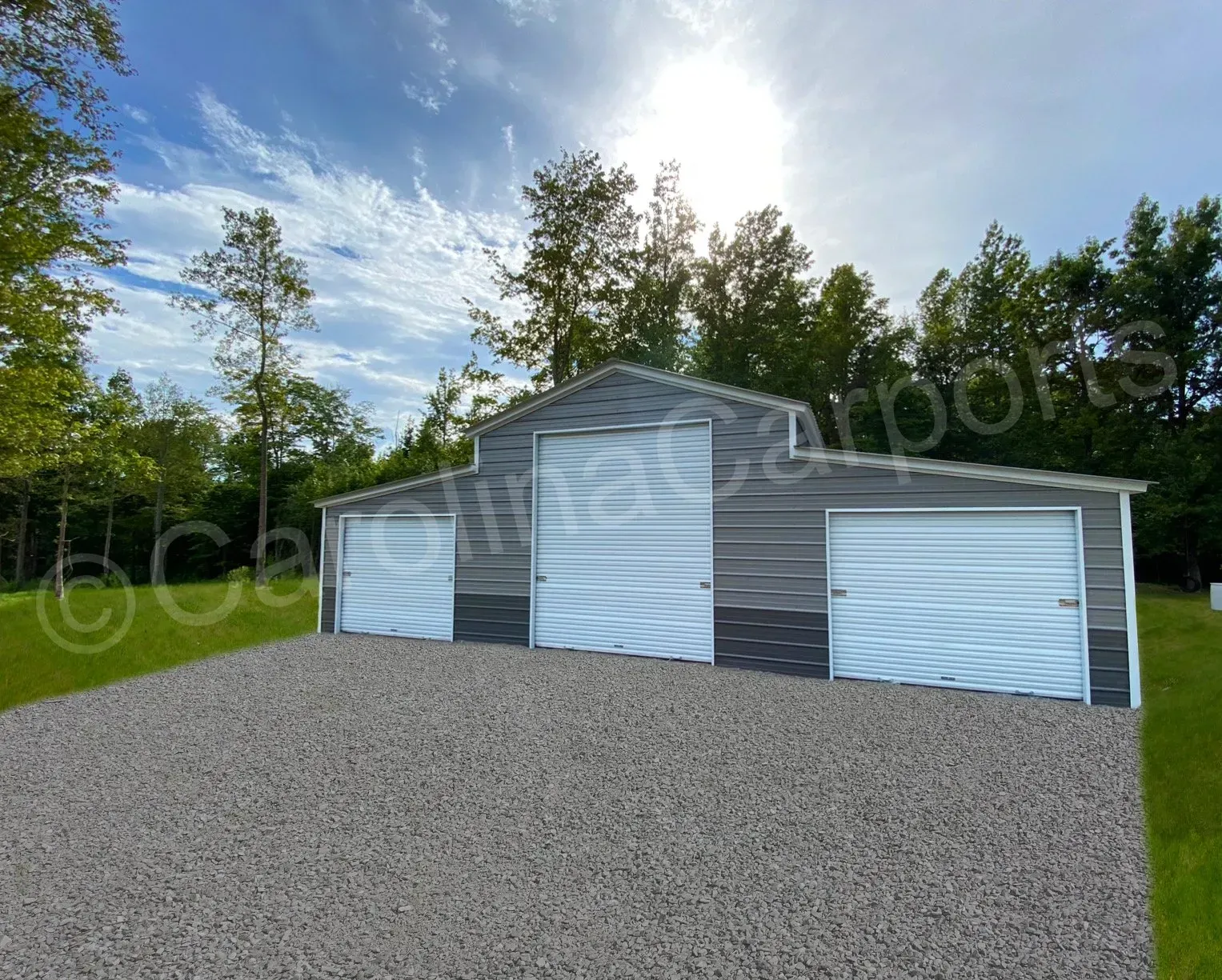 Steel gray and white metal barn with three garage doors on a gravel driveway, in front of trees.