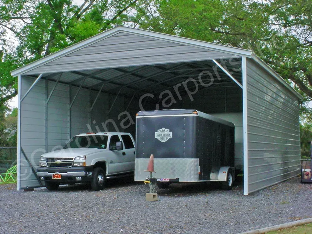 Gray metal carport sheltering a white pickup truck and a black trailer on gravel.