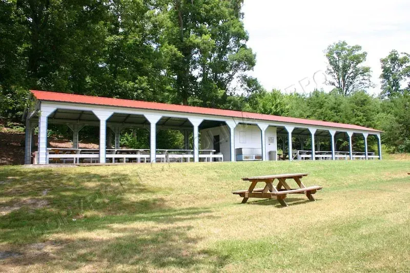 Long white picnic shelter with red roof, picnic table on grass, trees in background.