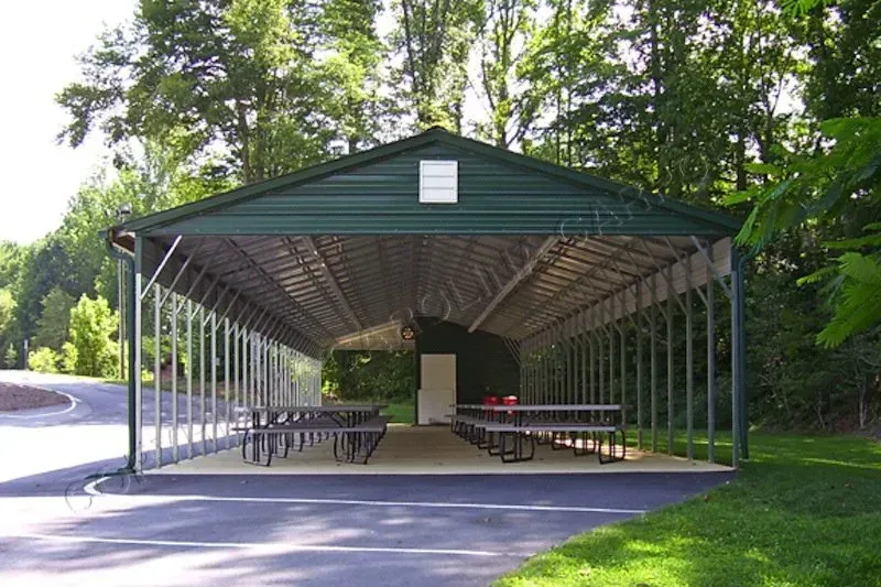Long, covered picnic shelter with tables, green roof, and forest background.