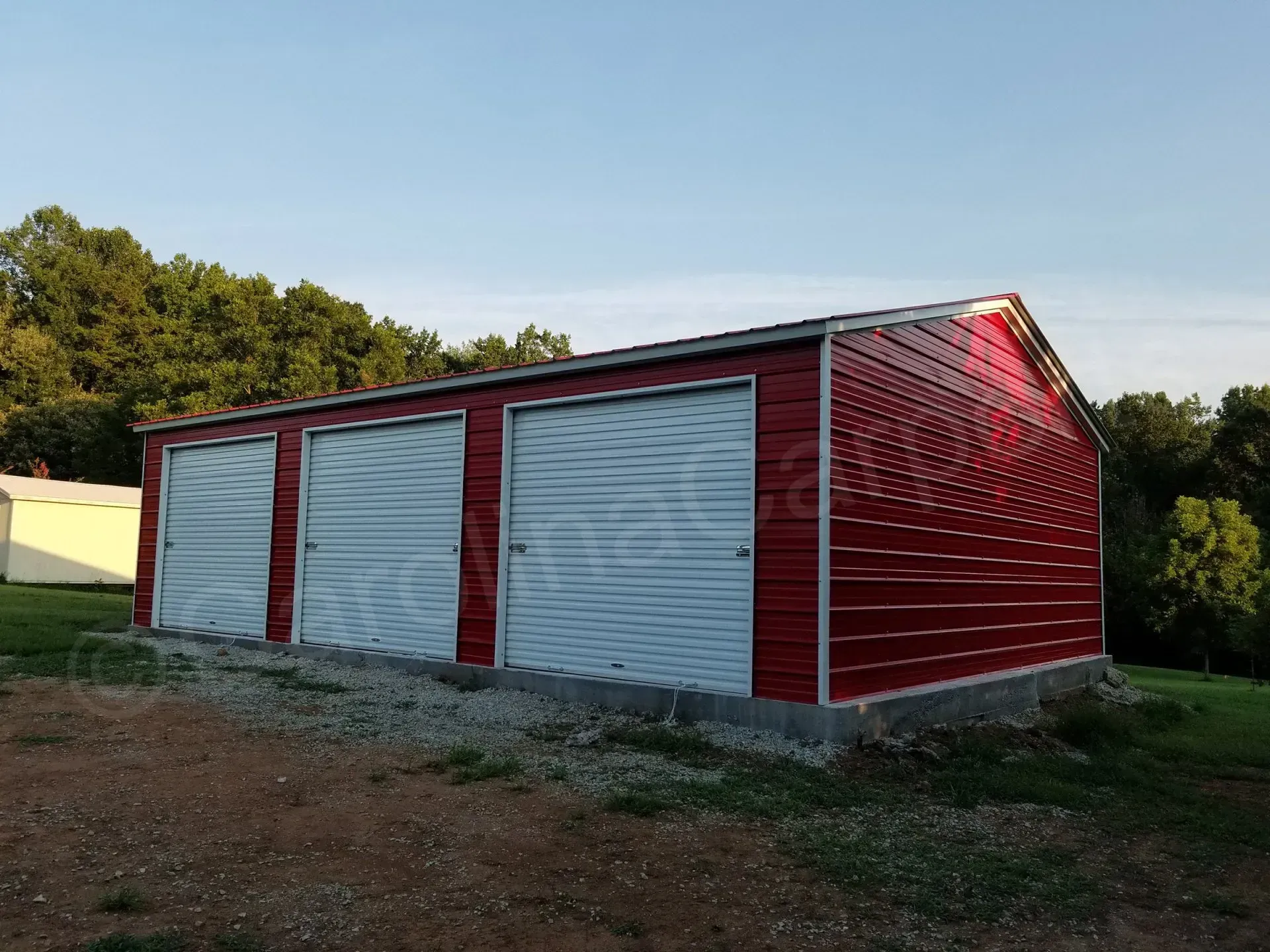 Red metal garage with three bays, light blue roll-up doors, and a gravel base.
