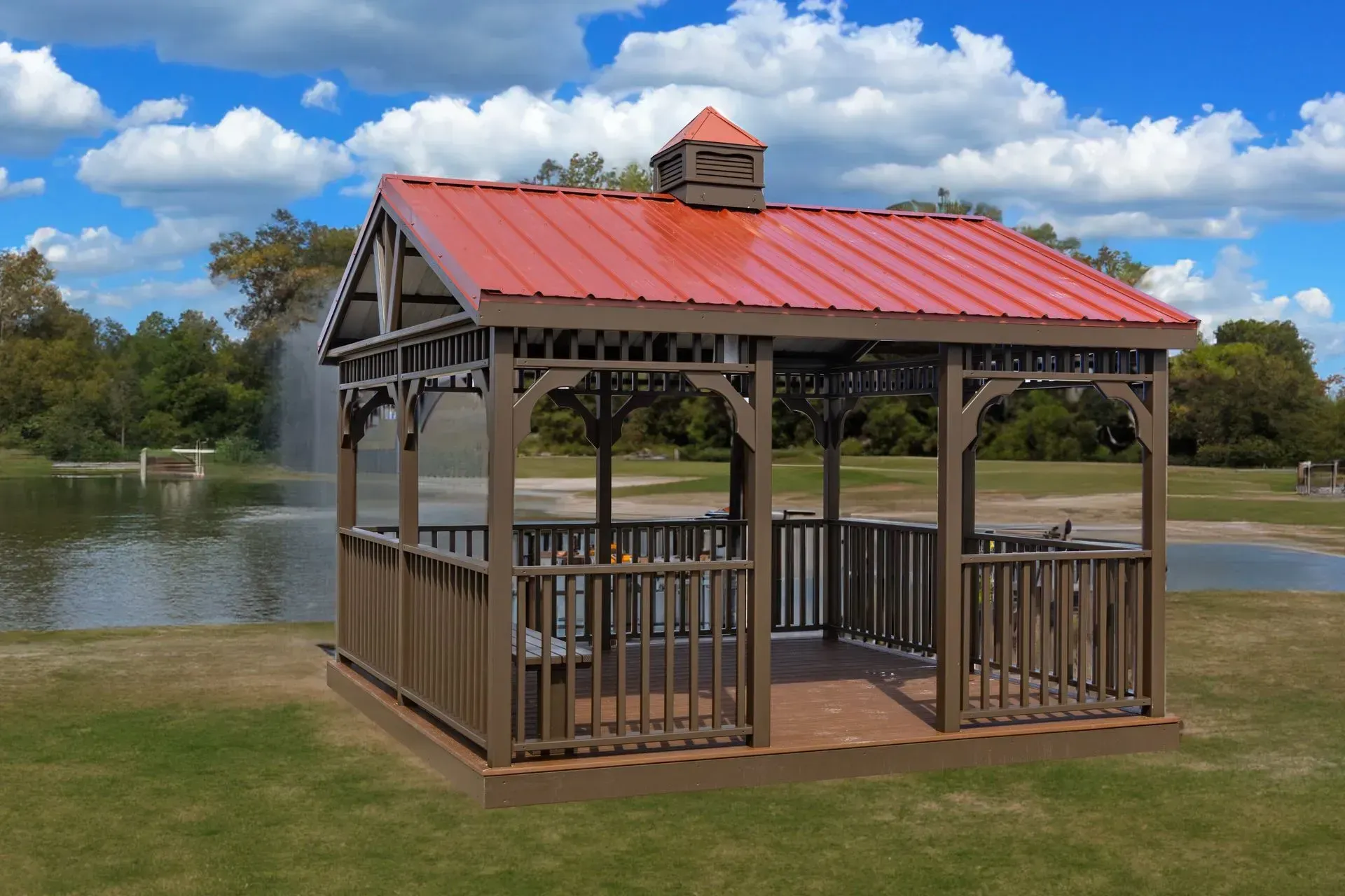 Brown gazebo with red roof on green grass by a lake with a fountain and blue sky.
