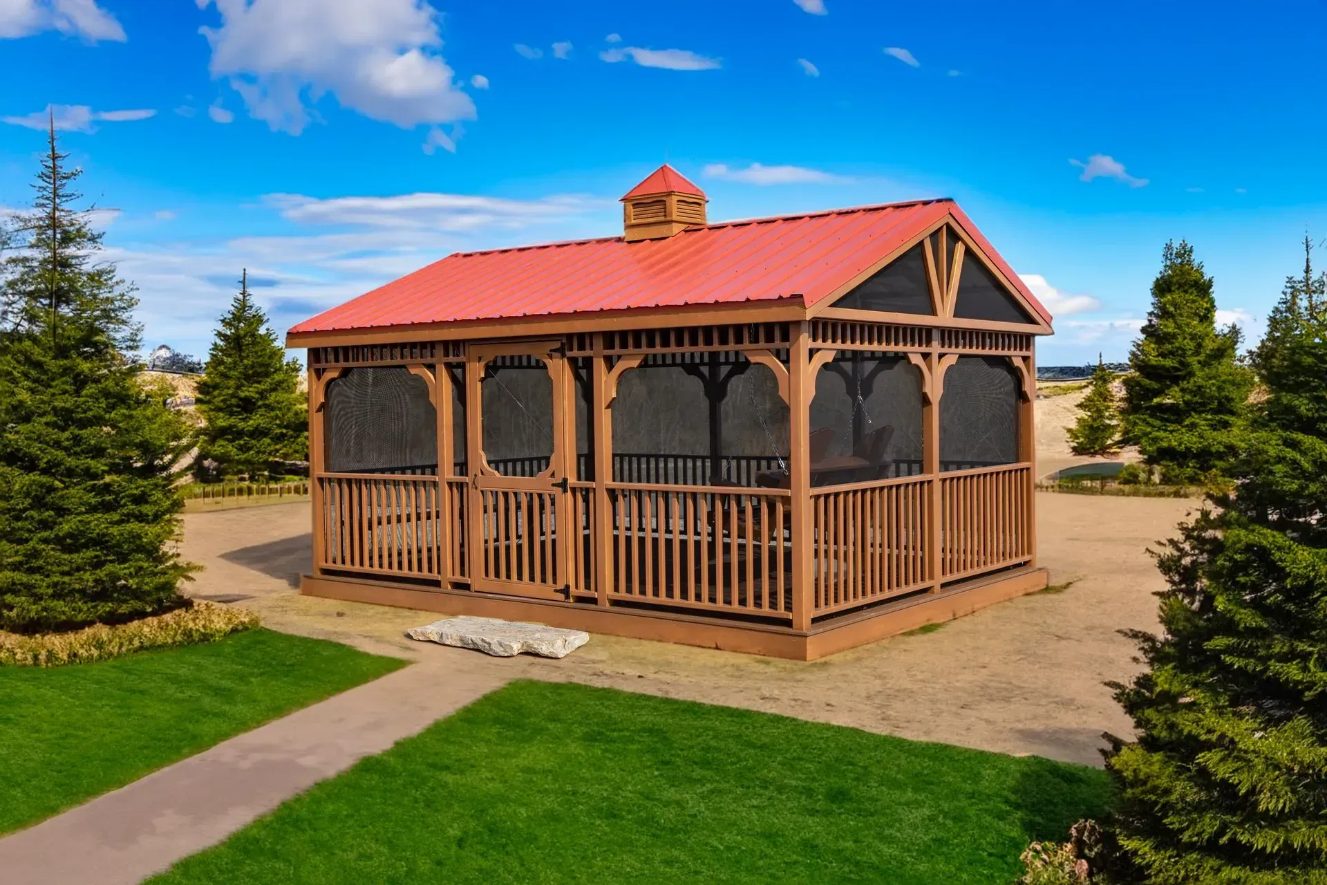 Wooden gazebo with red roof, screened walls, and brown railing, set on a gravel area with green lawn and trees.