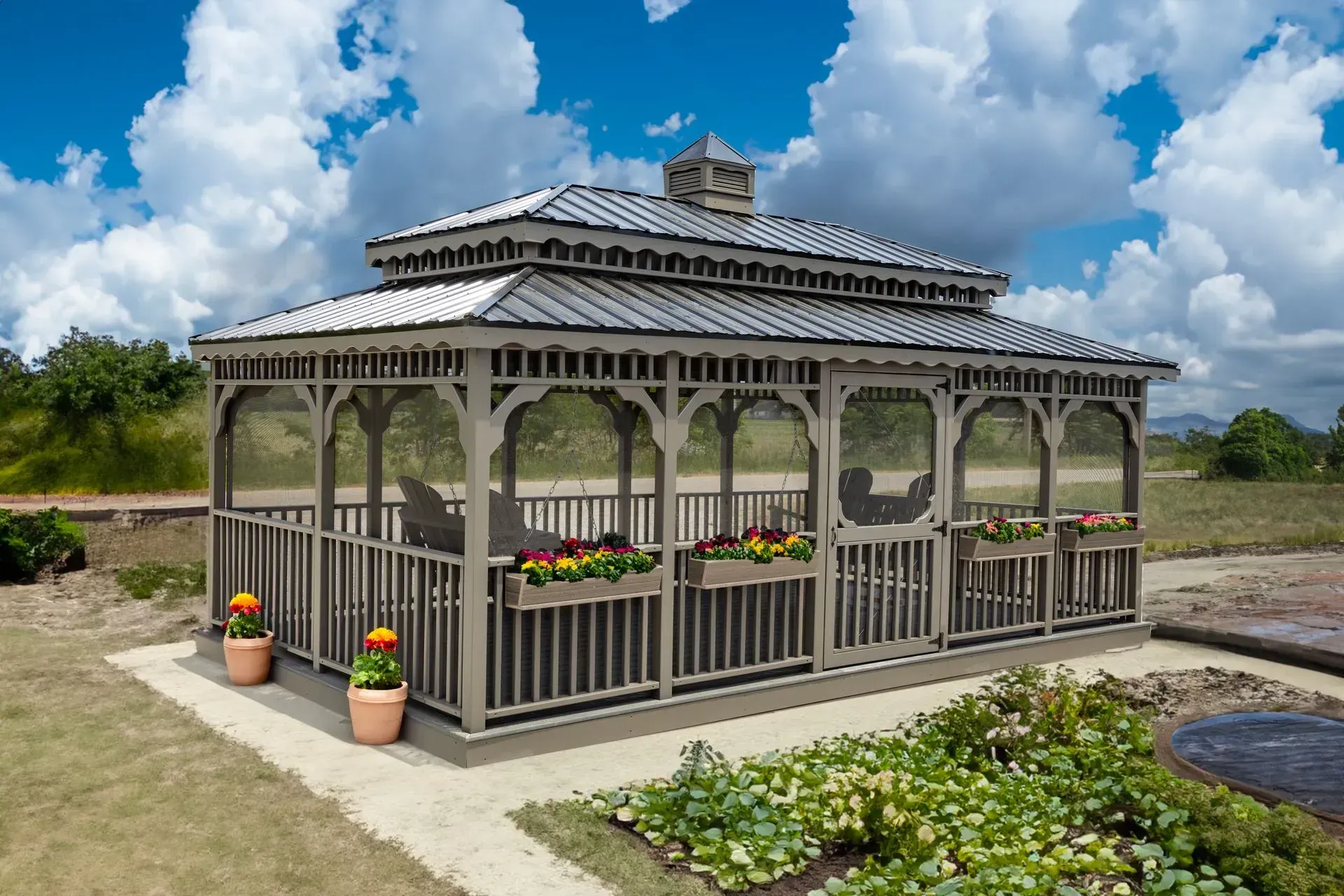 Gray gazebo with flower boxes on a concrete base, set in a garden with a cloudy sky.