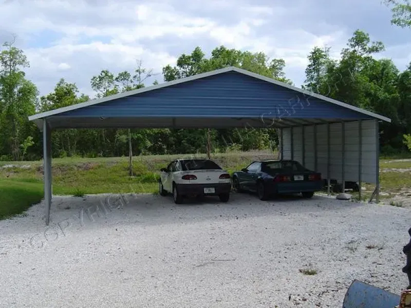 Two cars parked under a blue metal carport on a gravel surface. Trees and sky in the background.