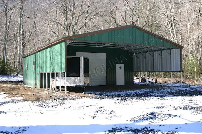 Green metal shed with an open-air carport, snowy ground, and trees in the background.