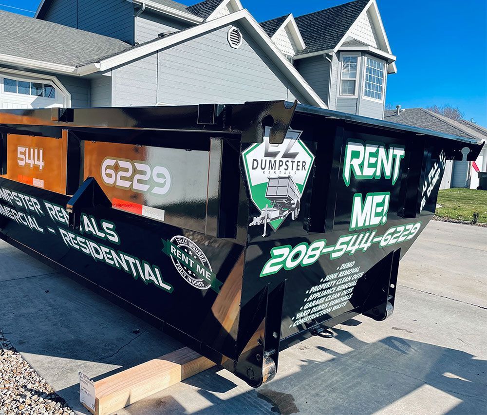 Black dumpster trailer with white/green “Rent Me” lettering parked by a house on a sunny street.
