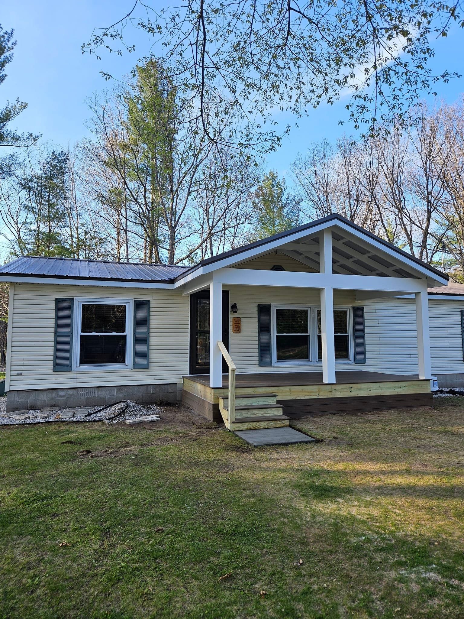 A single-story, light yellow house with dark shutters, a front porch with white pillars, and a metal roof in a wooded area.
