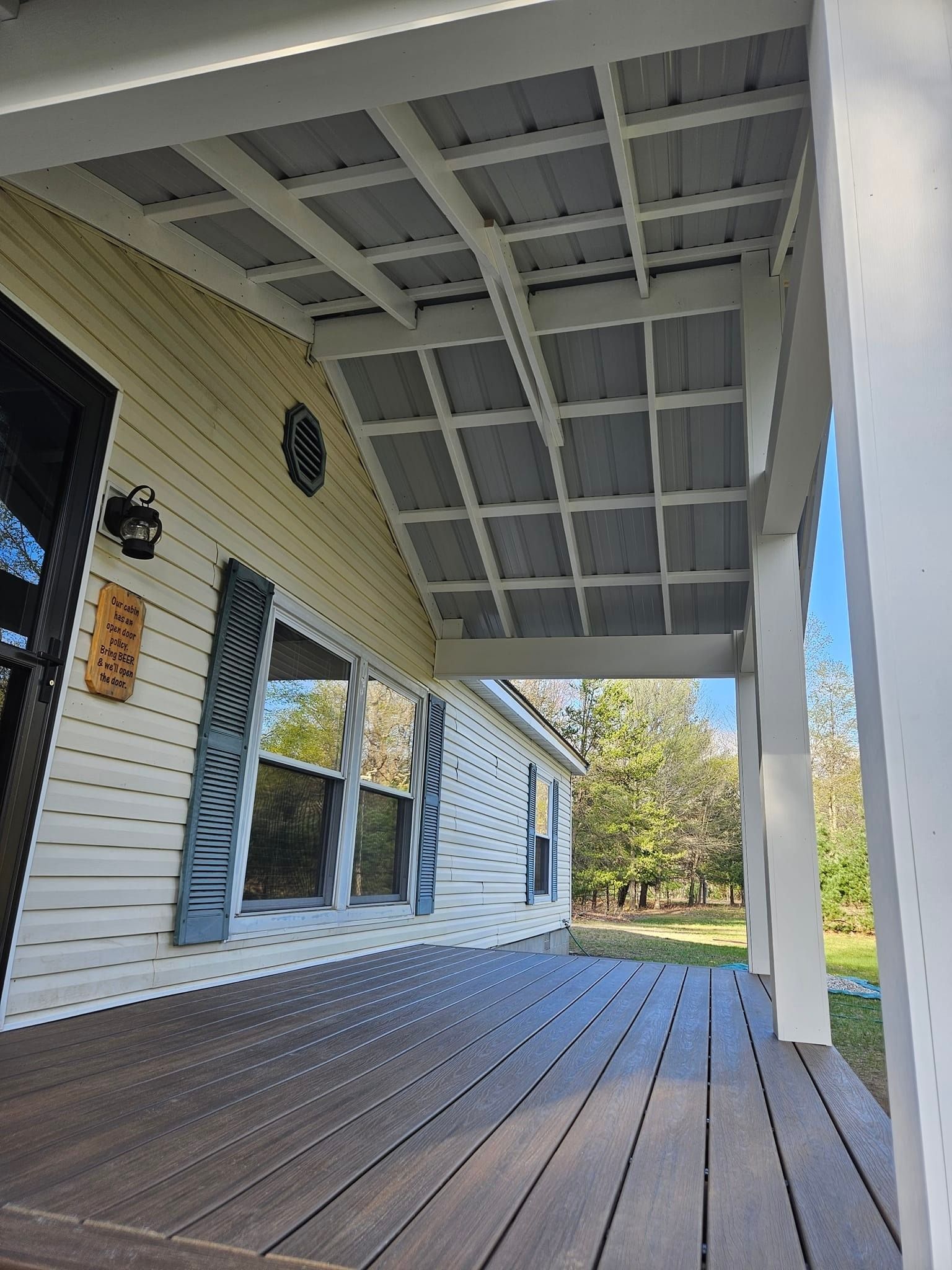 A low-angle view of a porch with brown wooden decking, light yellow siding, dark shutters, and a grey corrugated ceiling.