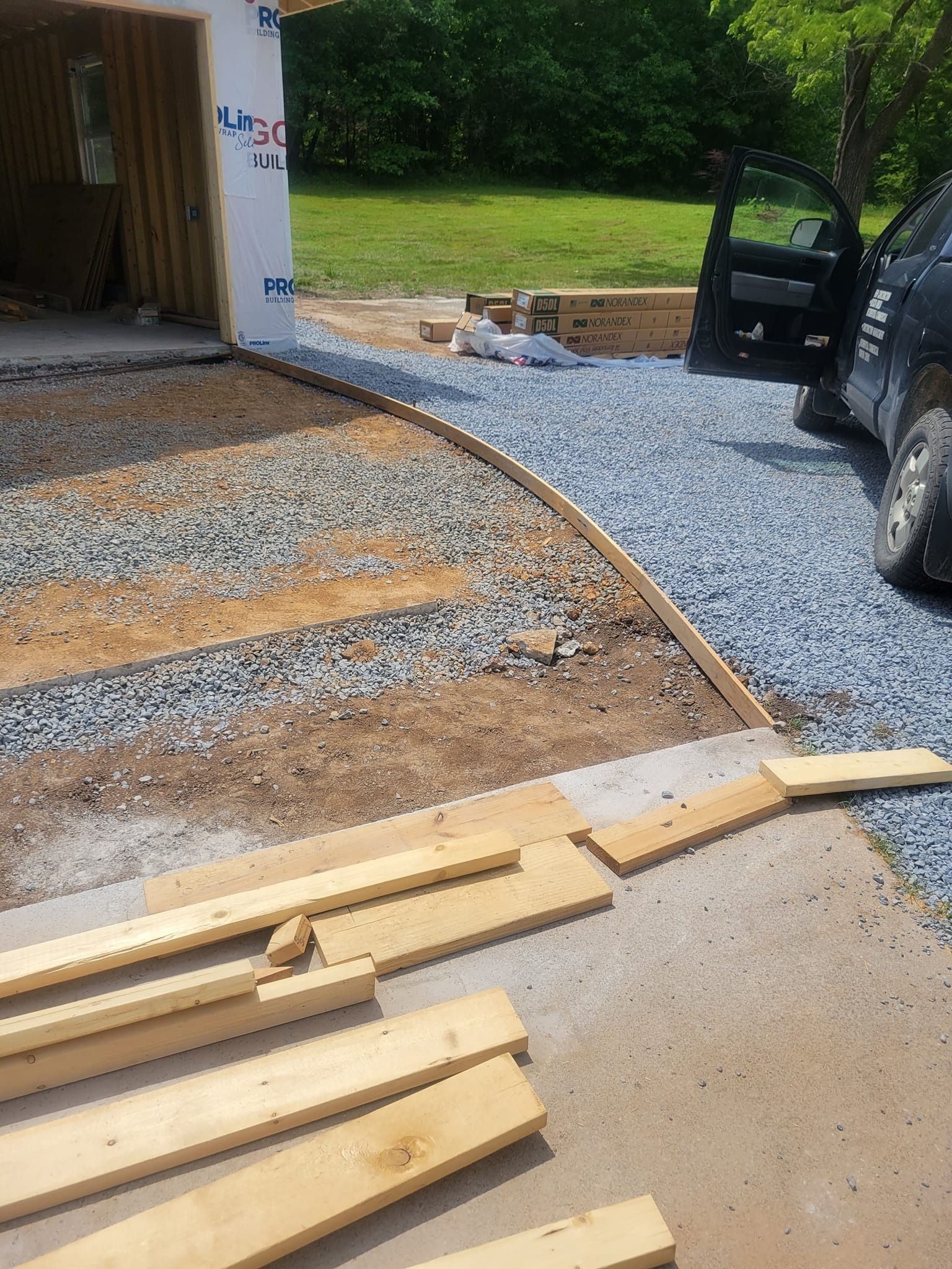Construction site showing a gravel driveway under preparation, with wooden boards and formwork framing the entrance.