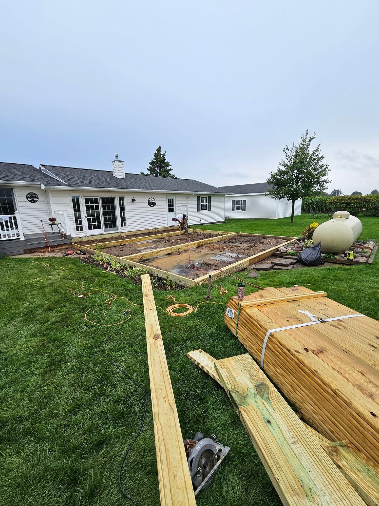 A backyard under construction with a wooden deck frame, stacked lumber, a circular saw, and a house in the background.