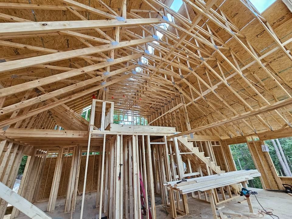 Framed house interior under construction with exposed wooden trusses, a loft platform, and partially built stairwell.