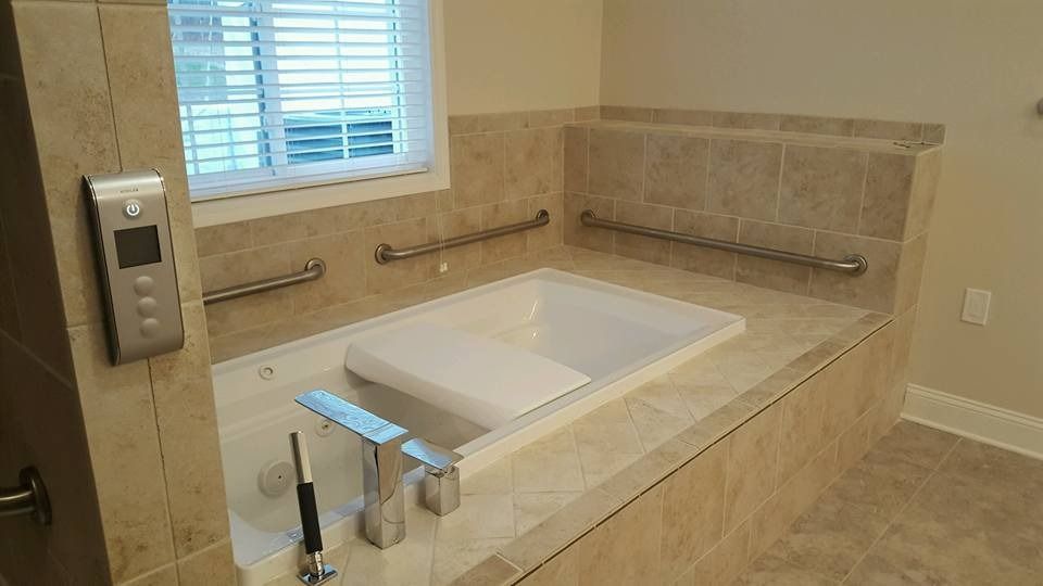A tiled soaking tub with grab bars, a built-in seat, and a digital control panel on the wall in a beige bathroom.