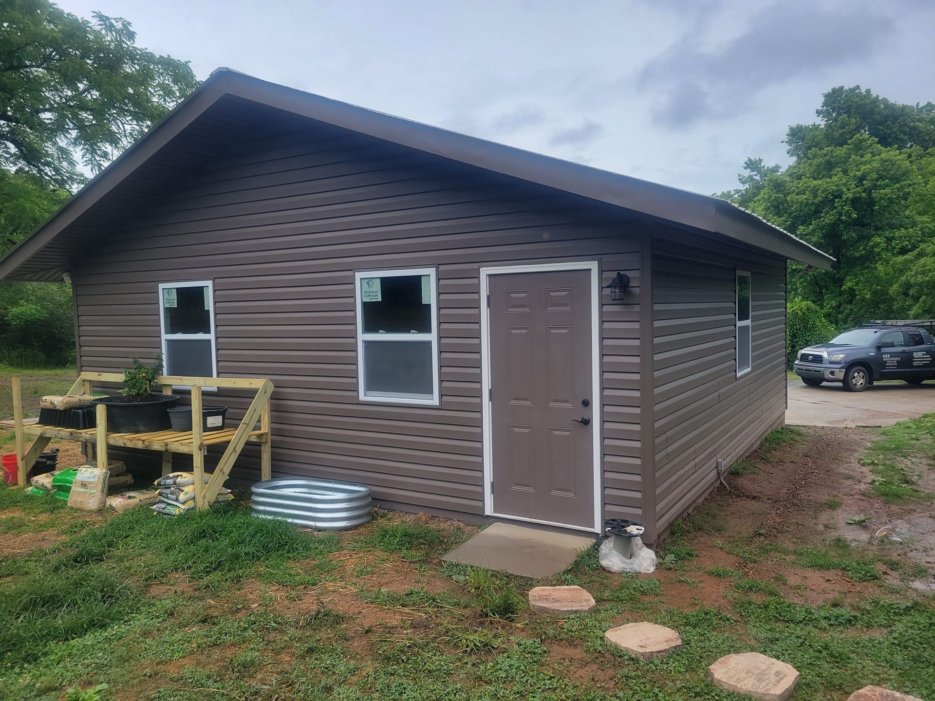 A brown shed with white-trimmed windows and a door, featuring a raised garden bed and a metal planter on a grassy lawn.