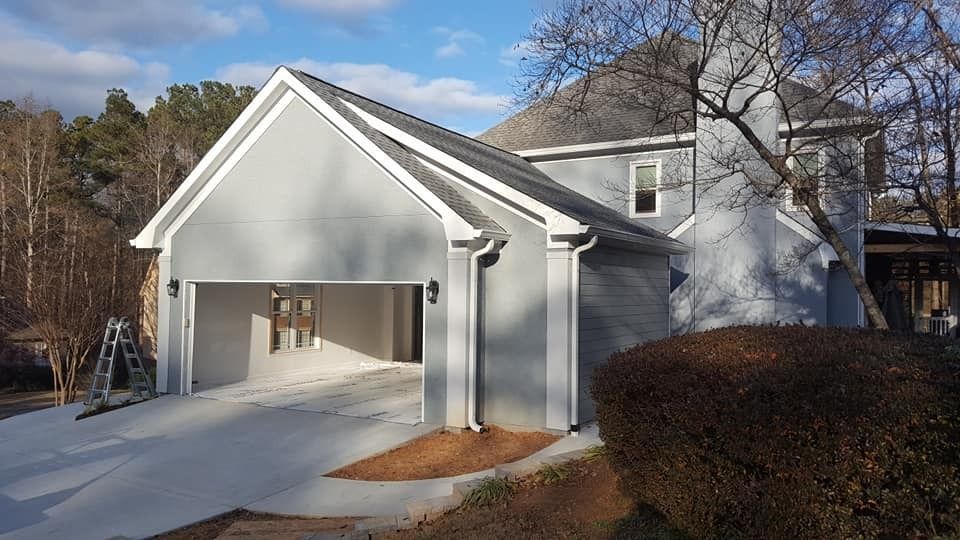 A light gray house with a prominent, open two-car garage, a concrete driveway, and a large bush in the foreground.