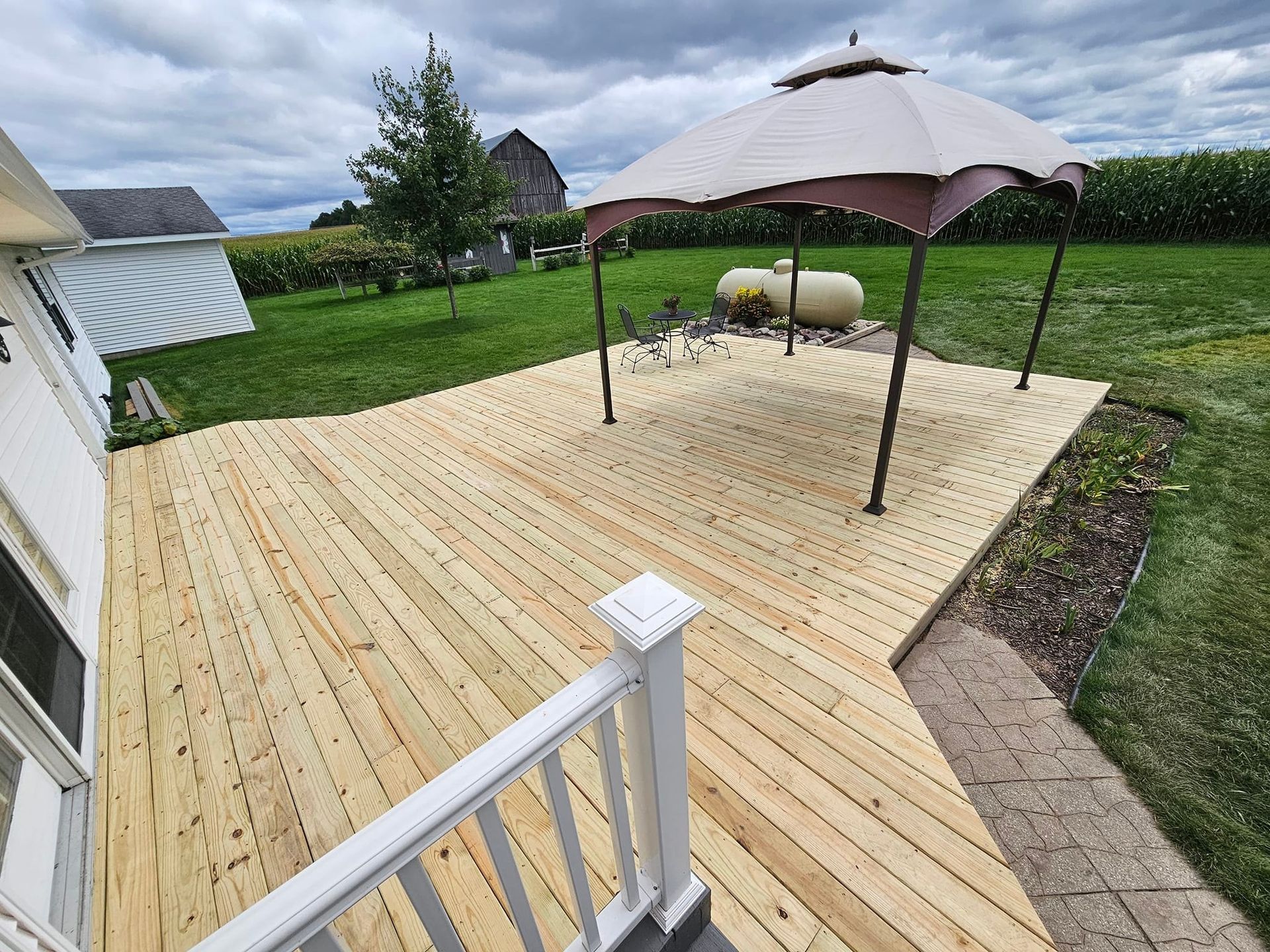 A new wooden deck in a backyard with a gazebo, white railings, and a view of a grassy field under a cloudy sky.