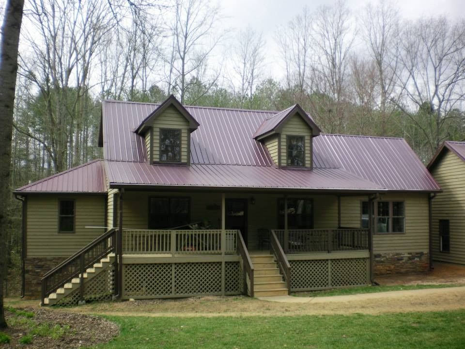 A two-story green cabin with a dark red metal roof, a large front deck with stairs, and two dormer windows in the woods.