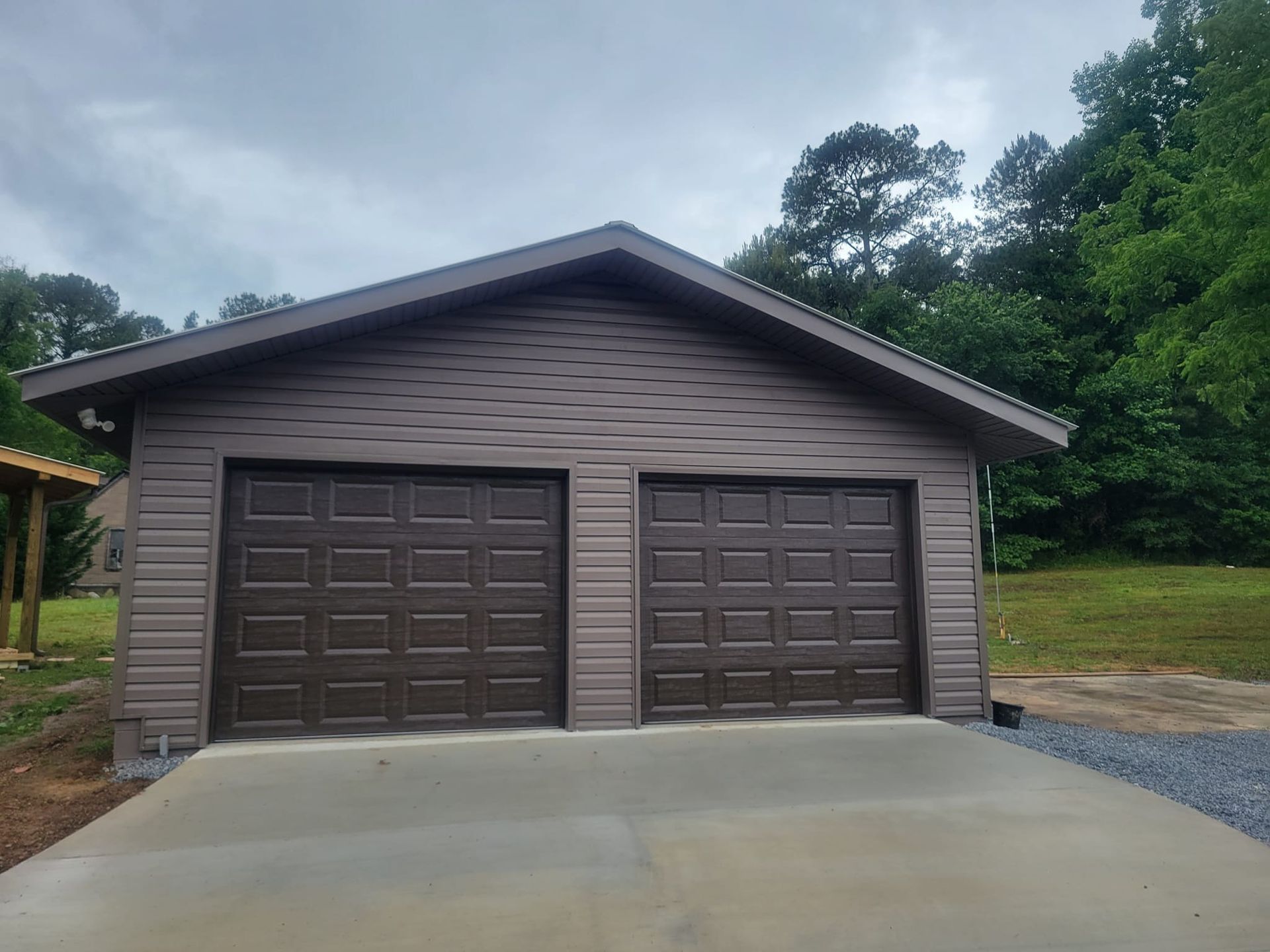 A detached two-car garage with brown siding and matching doors, situated in a wooded area with a concrete driveway.