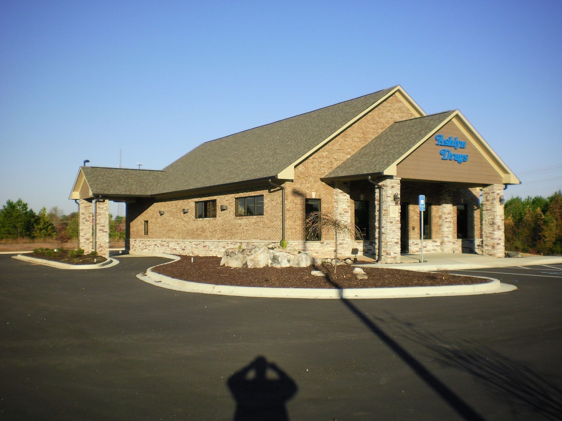 Tan brick building with a stone base and gray roof, featuring a small parking lot and a front entrance under a portico.