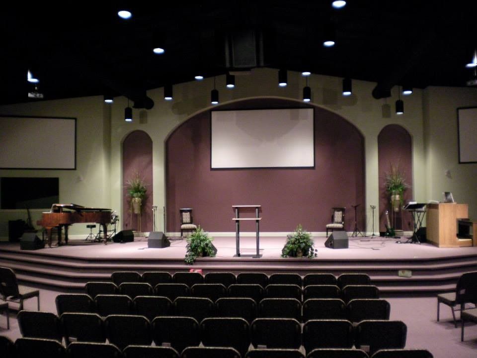 A stage with a central pulpit, two chairs, plants, and side screens, set in a dimly lit auditorium with theater seating.