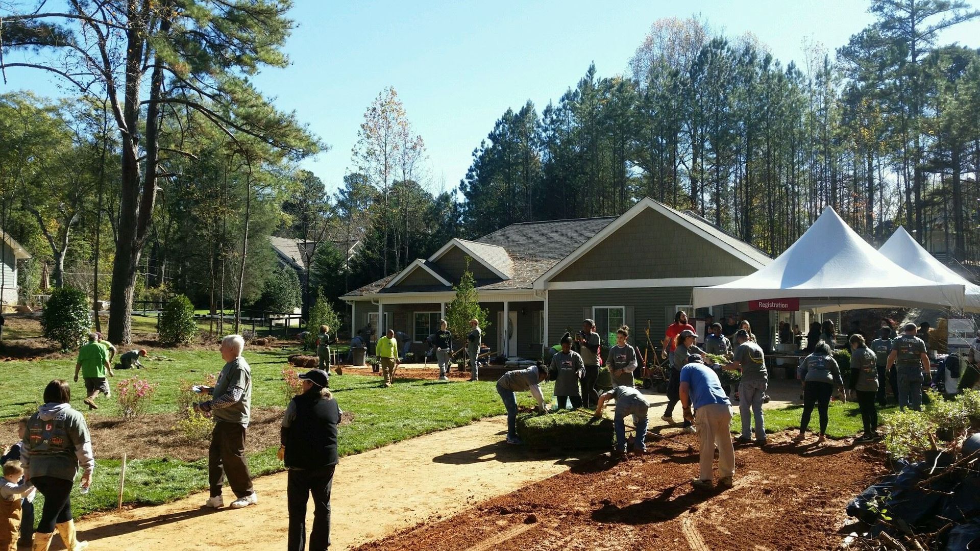 Volunteers work in a garden near a house with white tents on a sunny day.