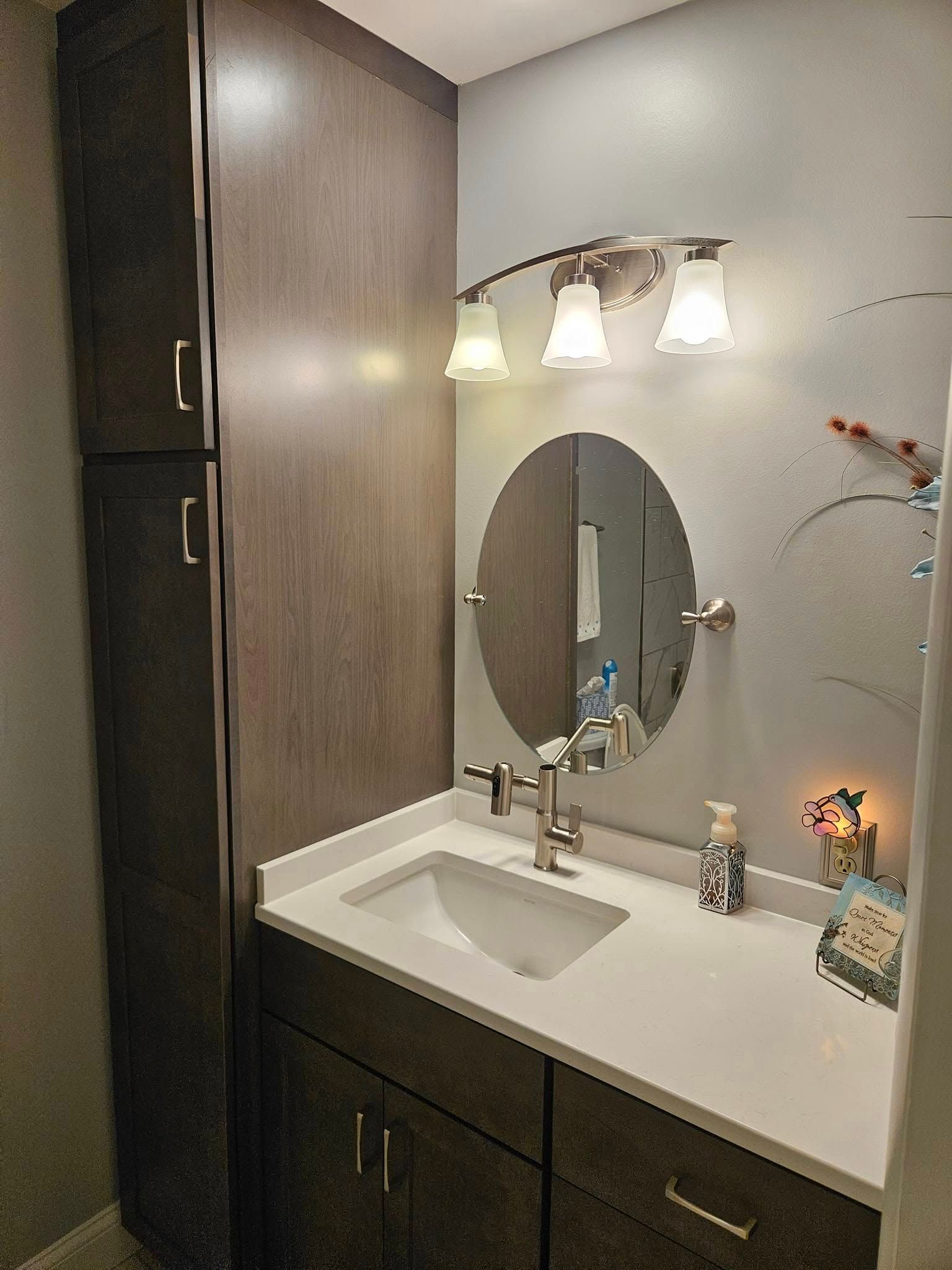 A modern bathroom vanity with dark wood cabinets, a white countertop, circular mirror, and three-bulb lighting fixture.