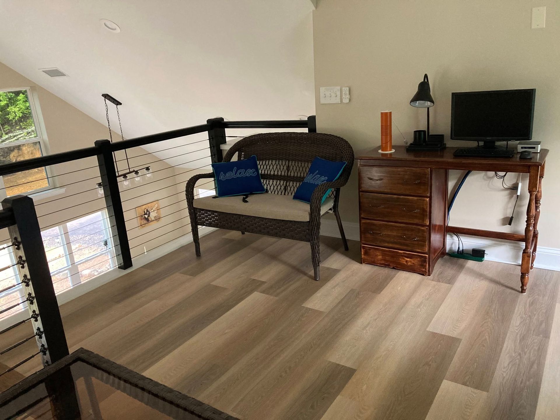 A loft space featuring a wicker loveseat with blue pillows, a wooden desk with a computer monitor, and light wood flooring.