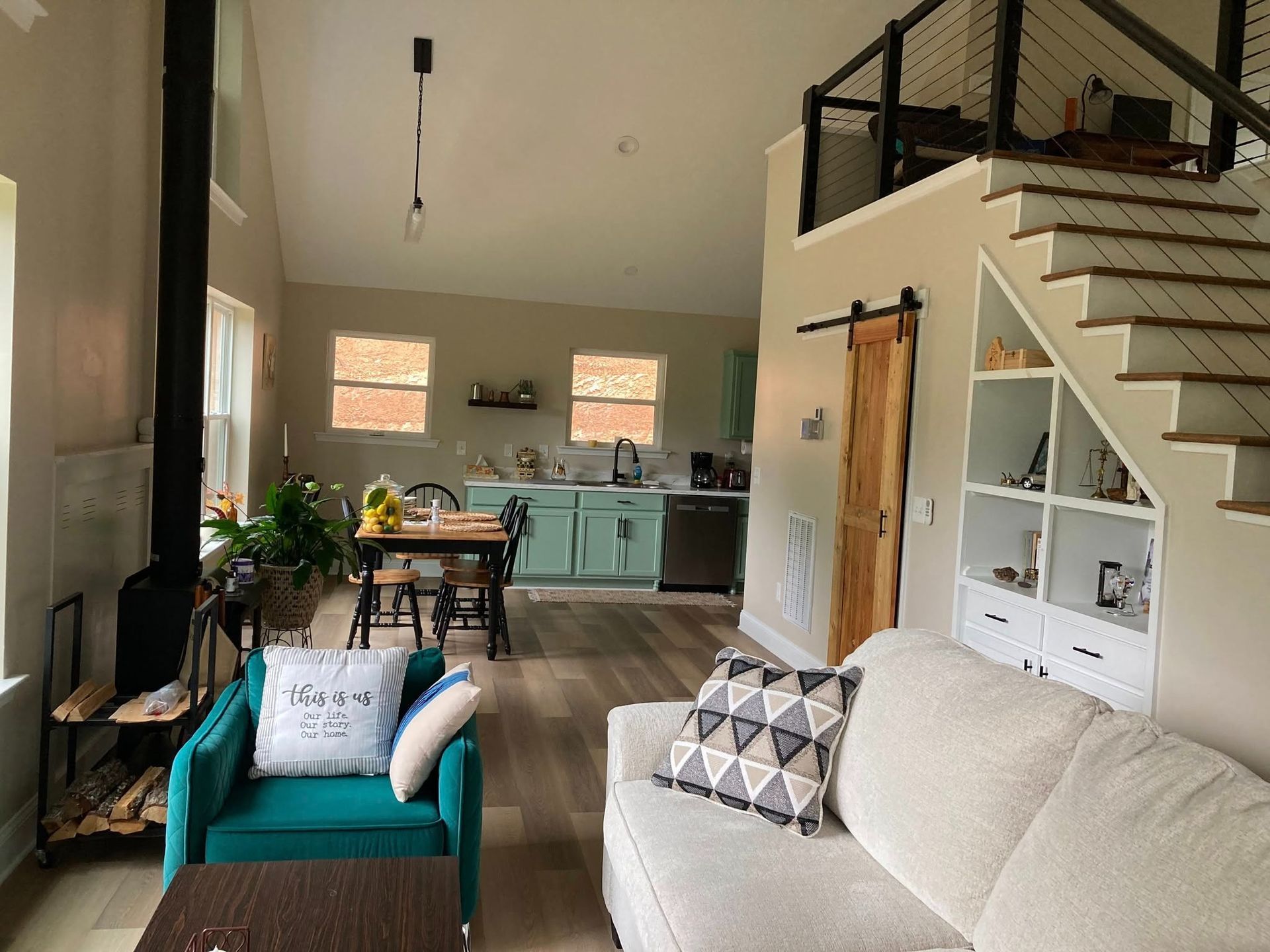 An open-plan living room and kitchen featuring a teal armchair, beige sofa, wooden dining table, and stairs with storage.