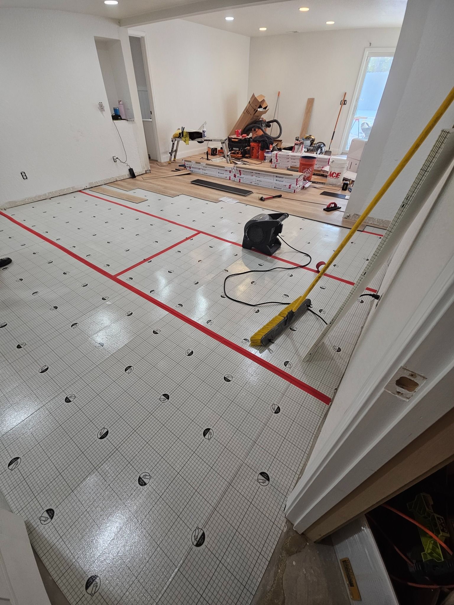 Radiant floor heating system installation with tubing grid on a white underlayment, floor tools, and room in progress.