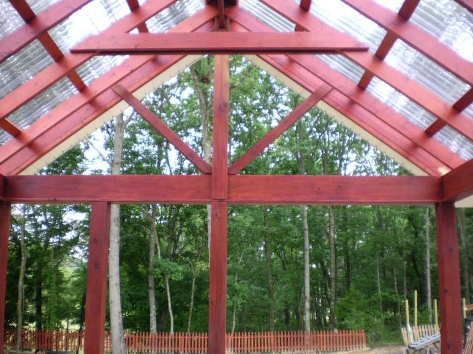 A wooden gazebo frame with a peaked roof and translucent roofing panels, set against a backdrop of trees.