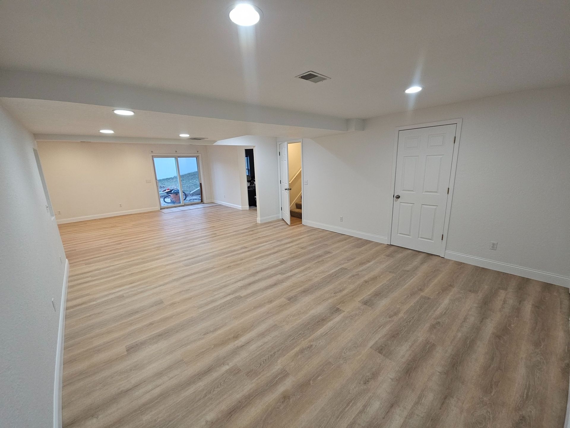 A large, open-concept room with light wood-look flooring, white walls, recessed lighting, and a white interior door.