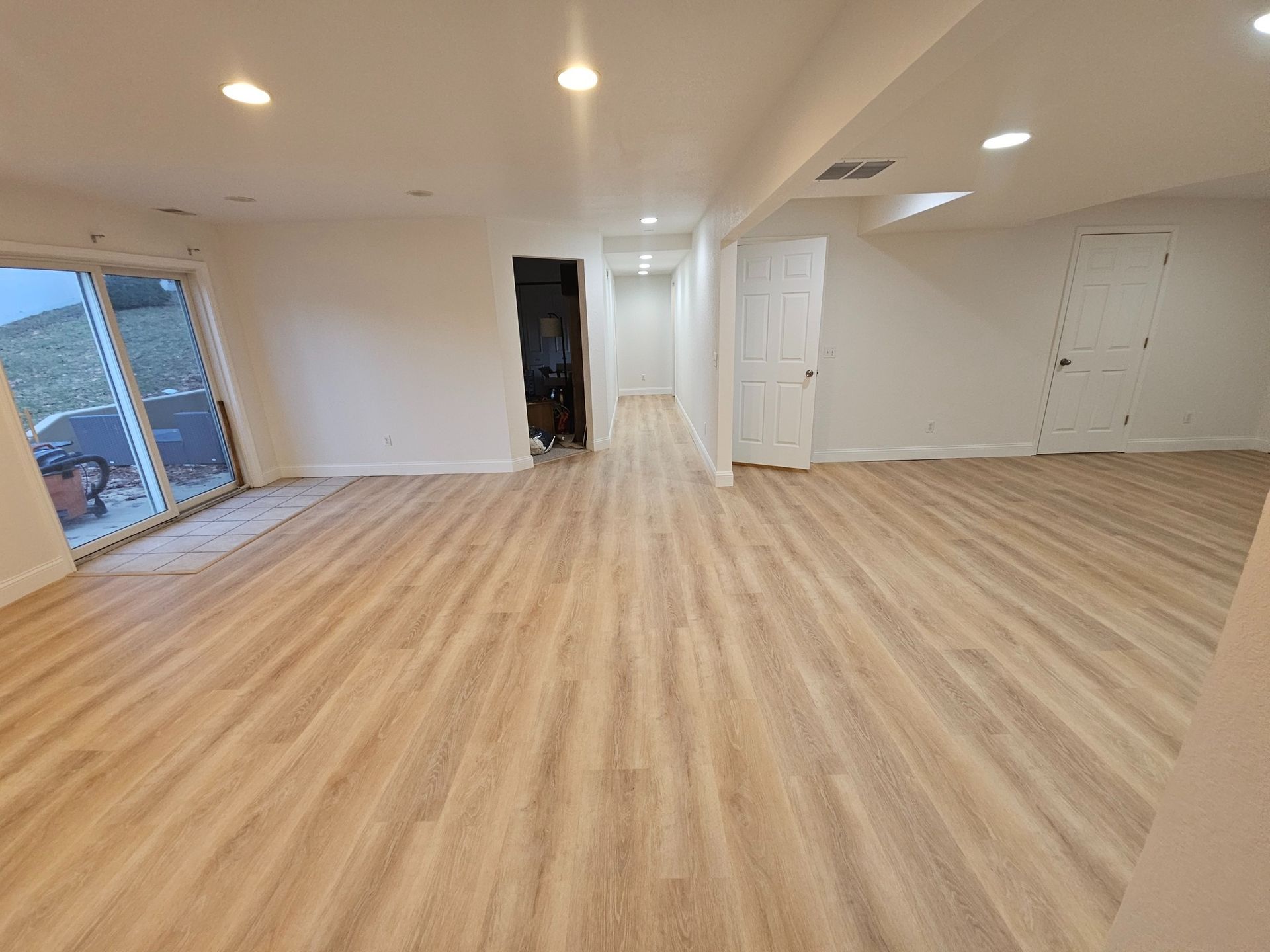 A wide, empty basement room featuring light wood-look flooring, white walls, recessed lighting, and two white doors.