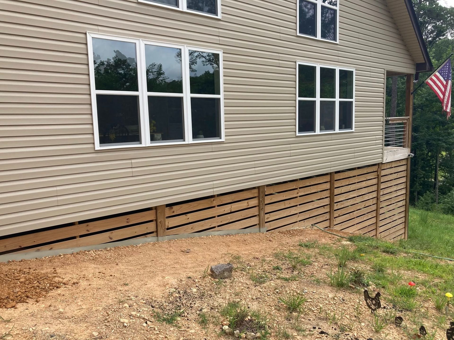 A side view of a house with beige siding, white-trimmed windows, a wooden lattice crawlspace cover, and an American flag.