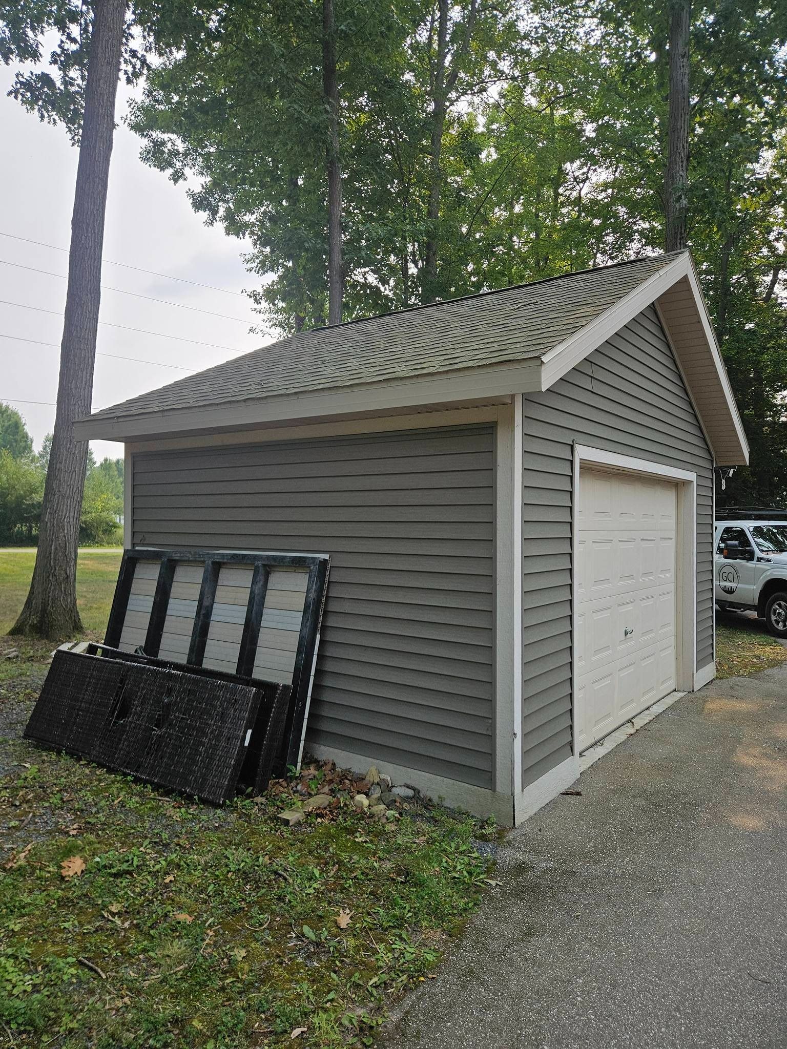 A gray detached garage with a white trim and door, set near trees with black panels leaning against the side.